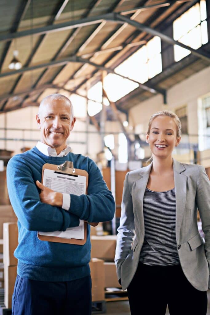 Two colleagues, a marketing director and an operations manager, stand side by side in a warehouse, smiling and looking proud. They are surrounded by shelves of goods and digital displays showing real-time data. The image represents their successful digital transformation efforts, optimizing warehouse operations and driving business growth.